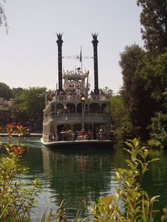 a large boat with people on it floating down a river next to trees and buildings