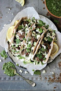 three tacos with meat, cheese and cilantro on a plate next to a bowl of pesto