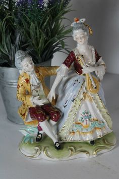 two figurines sitting next to each other near a potted plant