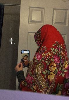 a woman in a red shawl taking a photo with her cell phone while wearing a headscarf