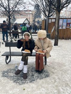 two people sitting on a bench in the snow eating hotdogs and drinking beer