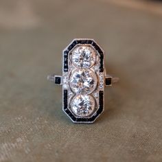 This antique Art Deco cocktail ring features 3.37 carats of Old European cut diamonds set in platinum and surrounded by pieces of square cut onyx. The three diamonds sit north-south along the finger in milgrain adorned bezel settings that appear to be floating inside their onyx frames. This ring is a great example of the fabulous angular style of the Art Deco era and is a perfect piece to accompany you to any cocktail party. Composition: Platinum Era: Art Deco 1915 - 1939 Current Ring Size: 8.50 Vintage Cocktail Rings Art Deco, Art Deco Cocktail Ring, Art Deco Three Stone Platinum Jewelry, Classic Diamond Ring With Black Diamonds, Art Deco Diamond Three Stone Ring, Art Deco Three Stone Diamond White Ring, Diamond White Three Stone Art Deco Ring, Classic Wedding Rings With Black Diamonds, Black Diamond Platinum Ring For Anniversary