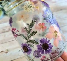 a hand holding a glass jar with flowers painted on the outside and inside, sitting on a wooden table