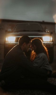 a couple sitting on the ground in front of a truck at night with their lights on