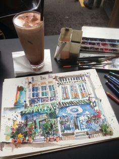 an open book sitting on top of a table next to a cup of coffee and watercolor pencils