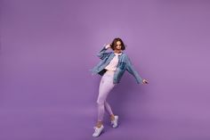 a woman in white pants and a denim jacket is posing for the camera on a purple background