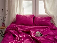 a cup of coffee sits on a pink bed in front of two windows with white drapes