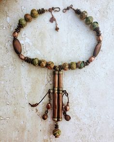 This necklace features an authentic vintage Tibetan Needle Box re-purposed into a piece of unique statement jewelry. I did not envision the piece to be overly colorful so I teamed up the copper patina on the pendant with the green, mustard brown and copper shades of the Kabamba Rhyolite Jasper stone beads. These beads are chunky and heavy - each one being 20mm wide so it gives the necklace a lot of oomph and character. The wood and copper accents add texture to the necklace and the Bronzite stone pebble beads tie the colors and ground the entire piece. I retained the authentic bells that came with the pendant (they hung on very worn out and frayed leather cords on either sides of the needle box) and restrung them in cow-hide leather cords along with copper accents to give the necklace move Bronze Hand Wrapped Bohemian Necklace, Rustic Patina Jewelry For Festival, Rustic One-of-a-kind Festival Necklaces, One Of A Kind Bronze Necklace For Festival, One Of A Kind Vintage Brown Jewelry, Unique Bronze Necklace For Festivals, Artisan Bronze Necklace For Festival, Bohemian Adjustable Patina Necklace, Unique One-of-a-kind Copper Necklace