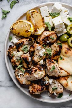 a white plate topped with chicken, potatoes and olives next to feta cheese