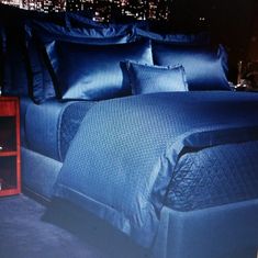 a bed with blue sheets and pillows in front of a cityscape at night