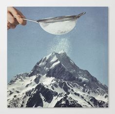 a hand holding a spoon over a mountain with snow on it's peak and the sky in the background