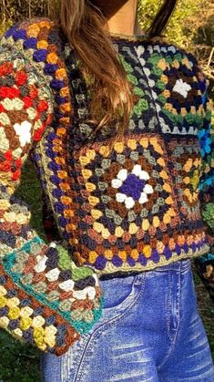 a woman wearing a multicolored crocheted sweater and jeans standing in the grass