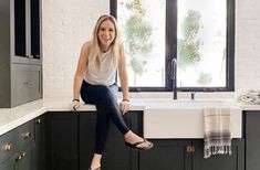 a woman sitting on the edge of a kitchen counter next to a sink and window