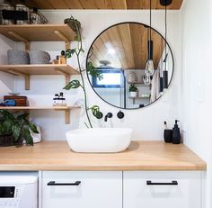 a white sink sitting under a round mirror on top of a wooden counter next to shelves