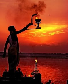 a man holding a lit oil lamp in front of a sunset over the water with other people