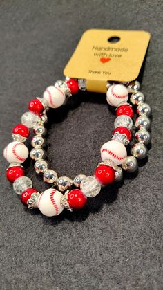 two red and white baseball bracelets with silver beads on a gray surface next to a yellow tag