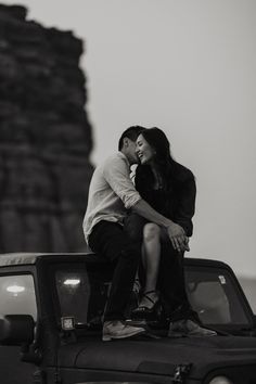 a man and woman sitting on the hood of a jeep in front of a rock formation