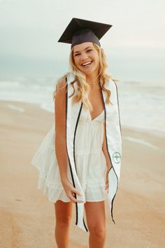 a woman in a graduation cap and gown standing on the beach with her hands in her pockets