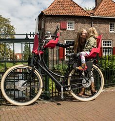 two women are sitting on a bicycle in front of a fence with a brick building behind them