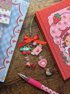 an open notebook with charms and a pink pen on a wooden table next to it