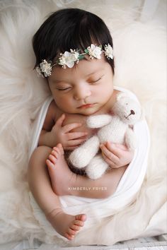 a baby is holding a teddy bear while wearing a flower headband and laying on a fluffy white blanket