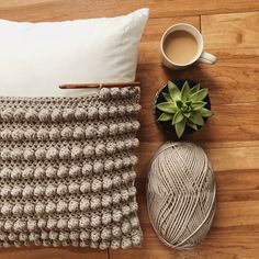 a crocheted pillow and yarn next to a cup of coffee on a wooden floor