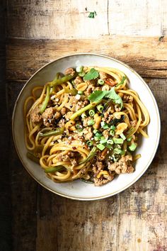 a white bowl filled with noodles and meat on top of a wooden table next to a fork