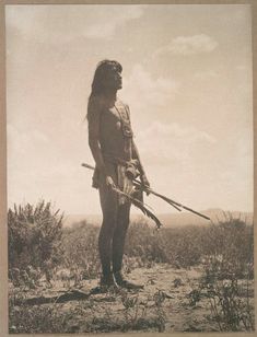 an old photo of a native american man holding two spears and standing in the desert