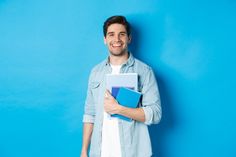 a young man is holding books and smiling at the camera while standing against a blue wall