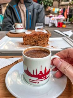 a person holding a cup of coffee in front of a slice of cake on a plate