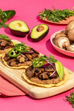 an assortment of food is displayed on a wooden cutting board and pink tablecloth with other foods