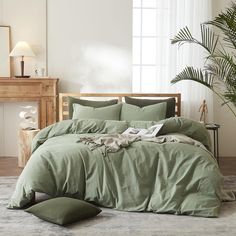 a bed with green sheets and pillows in a room next to a plant on the floor