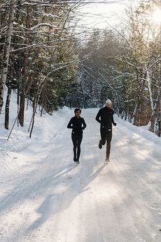 two people are running in the snow on a path between some trees and one person is wearing black