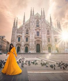 a woman in a yellow dress is standing near pigeons on the ground and buildings behind her