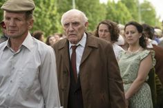 an older man wearing a brown jacket and tie walking with other people in the background