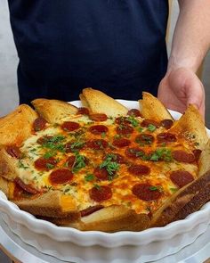 a person holding a pizza in a white dish on top of a wooden table next to bread sticks