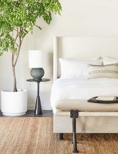 a bed with white linens and pillows in a bedroom next to a potted tree