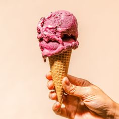 a hand is holding an ice cream cone with pink icing on it, while the other hand holds one