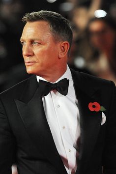 a man in a tuxedo looks off into the distance while wearing a red flower on his lapel