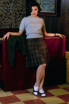 a woman standing next to a red couch wearing a plaid skirt and white shoes with her arms on the back of a chair