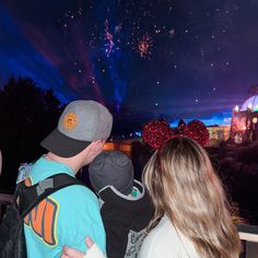 two people looking at fireworks in the sky with mickey mouse ears on their head and one person wearing a backpack