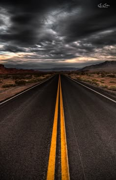 an empty road in the middle of nowhere with dark clouds above and yellow line painted on it