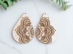 two wooden earrings with intricate designs on them sitting next to some green leaves and greenery