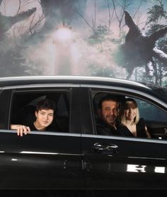 three people sitting in the passenger seat of a car with an image of dinosaurs on the wall behind them