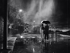 two people standing under an umbrella in the rain at night with buildings and street lights behind them