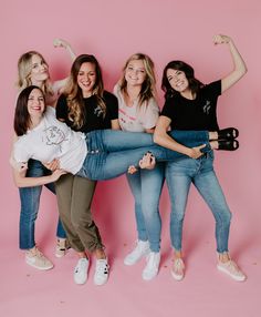 four women posing for a photo in front of a pink background with their arms up