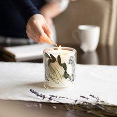 a person lighting a candle on a table with lavenders and coffee cups in the background