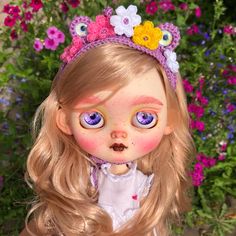 a close up of a doll with blonde hair and blue eyes wearing a flowered headband