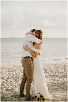 a bride and groom hug on the beach in front of the ocean during their wedding day