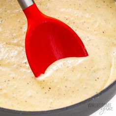 a red spatula in a pan filled with cheese and cream sauce on a white surface